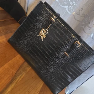 Svart datorväska med krokopräglad yta - Stilren svart datorväska med krokopräglad yta och guldfärgade detaljer. Väskan har flera fack, inklusive vadderat laptopfack med säkerhetsrem, och praktiska handtag. Perfekt för dig som vill ha en snygg och organiserad väska till skolan eller jobbet.