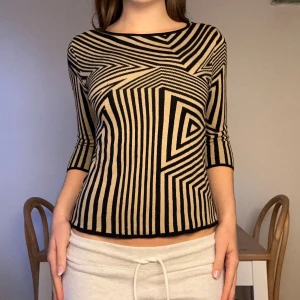 Beige och svart mönstrad topp - Säljer en beige och svart topp med grafiskt randigt mönster. Toppen har trekvartsärm och en rundad halsringning. Materialet är mjukt och stretchigt, perfekt för dig som gillar att sticka ut med coola mönster. Passformen är normal och toppen är riktigt bekväm.