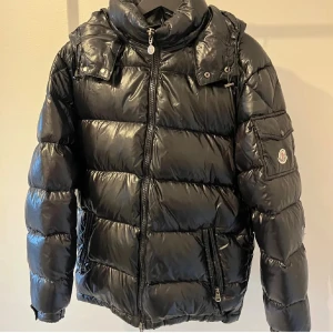 Svart Moncler pufferjacka med huva - Svart pufferjacka från Moncler med glansig finish, huva och dragkedja framtill. Jackan har en ficka med Moncler-logga på ärmen och flera praktiska fickor med dragkedja. Passformen är normal och jackan är perfekt för kalla dagar.
