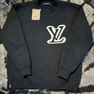 Svart stickad Louis Vuitton tröja - Svart stickad tröja från Louis Vuitton med stort vitt LV-logo på bröstet. Tröjan har rund hals, ribbade muddar och långärmad passform. Perfekt för dig som gillar exklusiva märken och vill ha en clean look med tydlig branding.
