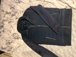 Mörkblå  superdry cardigan - Snygg mörkblå cardigan från superdry med quiltad framsida och stickade ärmar. Jackan har två dragkedjefickor och ribbade muddar vid ärmslut och nederkant. Perfekt för dig som gillar stilrent