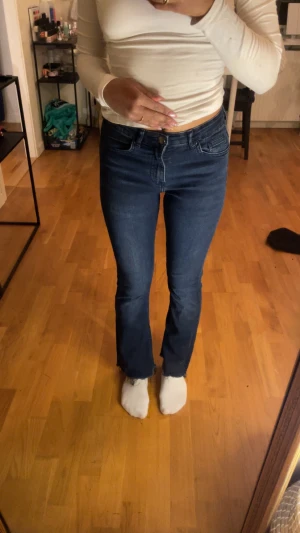 Blå bootcut jeans med rå kant - Snygga mörkblå jeans med bootcut passform och rå, fransad kant vid benslut. Klassisk femficksmodell med knapp och dragkedja framtill. Jeansen har en normal midja och är tillverkade i ett stretchigt denimtyg. Lite mindre i storlek Jeansen är använd i bra skick och säljs i befintlig skick. 