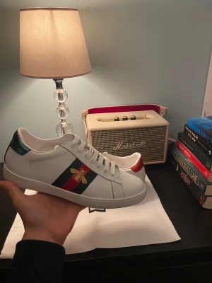 Gucci Ace sneakers med bi-detalj - Gucci Ace sneakers i vitt skinn med klassiska gröna och röda ränder på sidan och broderad guldfärgad bi. Baksidan har metallicröd och metallicgrön hälflik med textur. Platt sula med Gucci-logga och mönstrad undersida. Snygg och ikonisk modell för dig som gillar lyxiga detaljer.