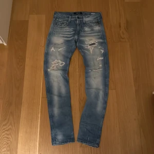 Replay Anbass ljusblå slitna jeans - Replay Anbass jeans i ljusblå tvätt med slitningar och trasiga detaljer framtill. Skinny passform med fem fickor och klassisk knappstängning. Snygga kontrastsömmar och Replay-logga på fickan. Perfekt för dig som gillar en edgy streetstil. Pris kan diskuteras😄