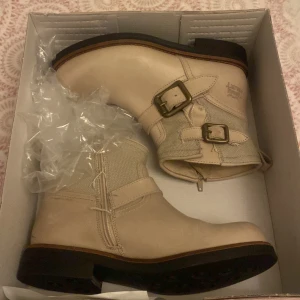 Beige boots från The Urban Project - Snygga beige boots från The Urban Project med robust platt sula och två coola spännen i metall på sidan. Skorna är tillverkade i skinn och canvas, har rund tå och detaljerade sömmar som ger en tuff look.