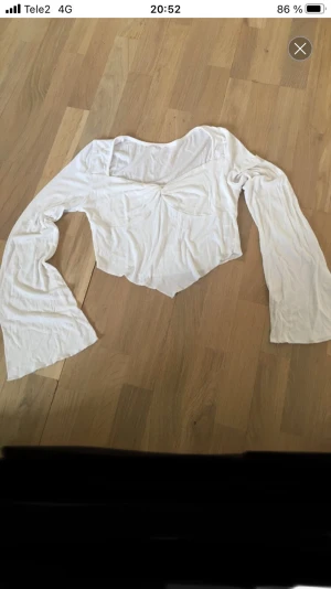 Vit croppad blus med trumpetärm - Säljer en vit blus med croppad passform och snygg twistad detalj framtill. Toppen har långa trumpetärmar och en v-ringad hals. Materialet är mjukt och lätt, perfekt för dig som gillar en chill och trendig stil.