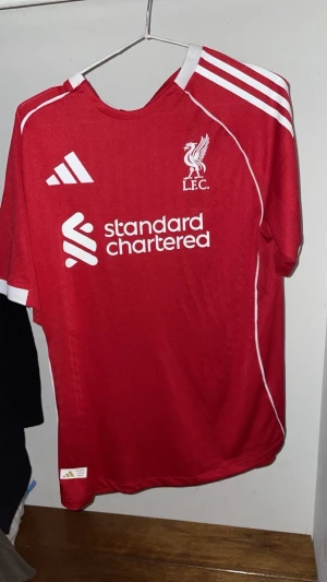 Liverpool FC röd matchtröja Adidas M - Liverpool FCs officiella matchtröja i rött från Adidas med vita detaljer och tre klassiska ränder på axlarna. Tröjan har klubbmärke och sponsortryck framtill, samt är tillverkad i ett lätt och ventilerande polyestermaterial. Slim fit-modell med korta ärmar.