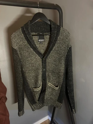 Svart och beige kofta från Smog - Slim fit kofta från Smog i svart och beige stickat material. Snyggt mönstrad med två framfickor och knappar hela vägen. Koftan har lång ärm och ribbade muddar vid ärmslut och nederkant. 