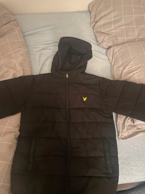 Svart pufferjacka från Lyle & Scott stl 160-164 - Svart pufferjacka från Lyle & Scott med huva och gul logga på bröstet. Jackan har dragkedja framtill, två sidofickor och quiltad design. Perfekt för kalla dagar och har en clean, stilren look som funkar till det mesta.