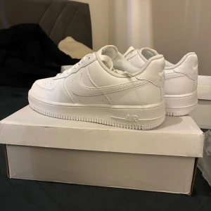 Nike Air Force 1 helvita sneakers - Säljer ett par klassiska Nike Air Force 1 sneakers i helvitt skinn. Modellen har låg profil, rund tå, snörning och den ikoniska swoosh-loggan på sidan. Perfekta för dig som vill ha en clean och tidlös look.