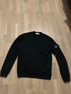 Svart Stone Island sweatshirt - Svart sweatshirt från Stone Island med klassisk rund hals och ribbade muddar. Tröjan har lång ärm och den ikoniska Stone Island patchen på vänster arm. Materialet känns mjukt och är troligtvis bomull eller polyester. Perfekt för dig som gillar streetwear och snygga detaljer. Använd några par gånger, men är i utmärkt skick. Pris kan diskuteras 