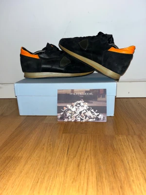 Philippe Model svarta sneakers med orange detalj - Svarta Philippe Model sneakers med snygga orange detaljer vid hälen. Skorna har rund tå, platt sula och snörning. Materialmix av skinn och mocka ger en cool och exklusiv känsla. Perfekt för dig som gillar stilrena sneakers med en twist.