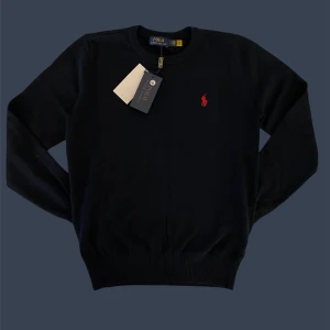 Mörkblå tröja från Polo Ralph Lauren - Snygg mörkblå tröja från Polo Ralph Lauren med klassisk rund hals och röd broderad logga på bröstet. Tröjan är långärmad och tillverkad i mjuk bomull, perfekt för en clean och stilren look.