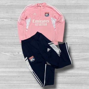 Olympique Lyonnais Tracksuit - Säljer en tracksuit med Olympique Lyonnais-logga och vita detaljer. Shortsen har resår i midjan och är tillverkade i ett lätt och svalt polyestermaterial, perfekta för fotbollsträning eller match.