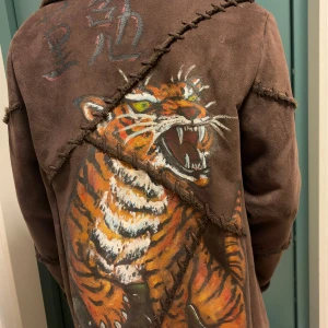 Brun mockajacka med handmålad tiger - Unik brun mockajacka med grova sömmar och handmålad tiger på ryggen. Jackan har knappar framtill, mjuk päls på insidan och krage. Snyggt detaljerad med asiatiska tecken och råa kanter som ger en cool vibe. Jag är S på bilden 178cm