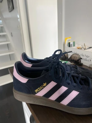 Adidas Spezial sneakers marinblå/rosa - Säljer ett par Adidas Spezial sneakers i marinblå mocka med ljusrosa detaljer och klassiska tre ränder på sidorna. Skorna har snörning, gummisula och guldfärgad Spezial-logga. Dem är endast använda fåtalet gånger men är i nyskick! Storlek 38 2/3. Skriv till mig om ni vill diskutera priset! 