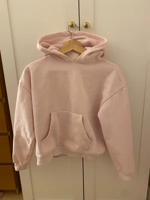 Ljusrosa hoodie med huva - Säljer en ljusrosa hoodie med stor känguruficka framtill och ribbade muddar vid ärmslut och nederkant. Tröjan har en skön huva och är perfekt för chill dagar. Passar dig som gillar en loose fit och enkel stil.