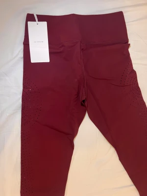 Vinröda leggings från La Nikar - Snygga vinröda leggings från La Nikar med hög midja och coola perforerade detaljer längs benen. Stretchigt syntetmaterial som sitter tajt och bekvämt. Perfekta för träning eller chill, med en modern look som sticker ut.