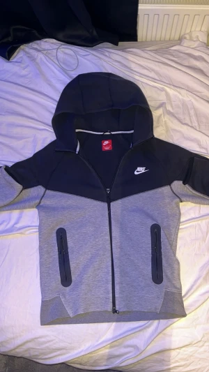 Nike svart och grå hoodiejacka - Nike tech i svart och ljusgrått med dragkedja hela vägen, två fickor med dragkedja och stor huva. Snyggt sportig look med logga på bröstet och kontrastpaneler. Perfekt för chill eller när du är på språng.