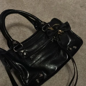 Svart handväska med rosett och hjärtdetalj - Snygg svart handväska i glansigt skinnliknande material med silverfärgade nitar och dragkedja framtill. Väskan har dubbla handtag, axelrem och en dekorativ svart rosett med guldfärgad hjärtdetalj som hänger framtill. Perfekt för dig som gillar edgy och söt stil.