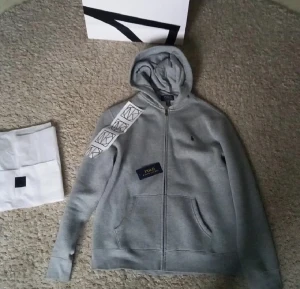 Ralph Lauren hoodie från NK - Säljer en stilren grå hoodie från Polo Ralph Lauren med dragkedja och broderad logga på bröstet. Den har en klassisk design med långa ärmar och en bekväm passform. Perfekt för en avslappnad look. Helt Ny från NK säljer då jag glömde av den och nu är den för liten för mig. Tar inte emot returer. Pris kan diskuteras vis snabbt köp. Tröjan är XL för barn och S för Män
