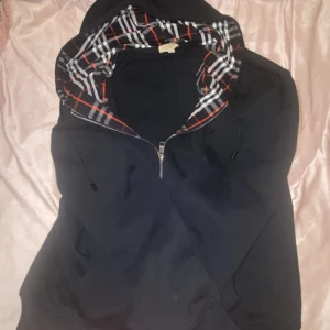 Svart Burberry hoodie med rutig huva - Svart hoodie från Burberry med klassisk rutig insida på huvan i vitt, rött och grått. Tröjan har snörning med metalländar, dragkedja framtill och broderad logga på bröstet. Perfekt för dig som gillar stilrena streetwear-plagg med lyxig touch.