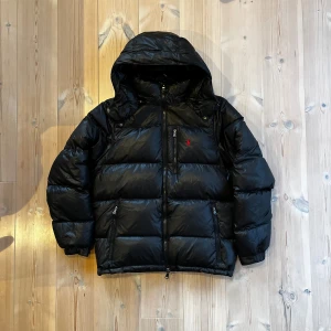 Svart Glansig Ralph Lauren Dunjacka - Svart glansig pufferjacka av dun från Ralph Lauren🪶 || Perfekt jacka för att hålla dig varm nu när det börjar bli som kallast!❄️ || Jackan är märkt med ”RL” på alla dragkedjor och det finns även en kardborre mudd som gör så ingen vind kommer in vilket håller dig varmare💨 || Huvan går även enkelt och snabbt att dra av med dragkedja || Jackan har endast en liten defekt och det är ett litet hål i fickan på vänster sida men det är ingen man ser eller knappt märker || Hör gärna av er vid fler frågor!🏌️