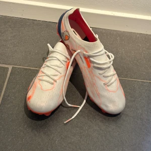 Puma Ultra Ultimate FG/AG “Lunar Pack” (limited edition) - Säljer ett par puma fotbollsskor i vitt med orangea detaljer och coola mönster. Skorna har snörning, platt sula med dobbar i orange och en unik innersula med astronautmotiv. Perfekta för dig som vill sticka ut på planen. En av dem få kvar så man får skynda sig haha.