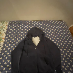 Mörkblå hoodie från Polo Ralph Lauren - Snygg mörkblå hoodie från Polo Ralph Lauren med dragkedja och huva. Tröjan har snörning vid huvan och en liten röd logga broderad på bröstet. Perfekt för chill dagar och enkel att matcha med jeans eller mjukisbyxor. Storlek M men passar även bra som S. Skick 9/10 möjligen en och annan tråd som är lös annars är skicket galant. Kom gärna med frågor och funderingar!!