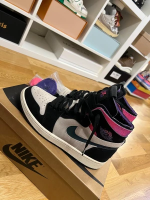 Nike Air Jordan 1 PSG Highs - Nike Air Jordan 1 Paris Saint-Germain high-top sneakers med svart och grå mocka, detaljer i rosa och lila samt PSG-logga på hälen. Swoosh i mörkblått och perforerad tå. Medföljer extra skosnören i lila och rosa. Riktigt snygga och unika sneakers för dig som vill sticka ut.