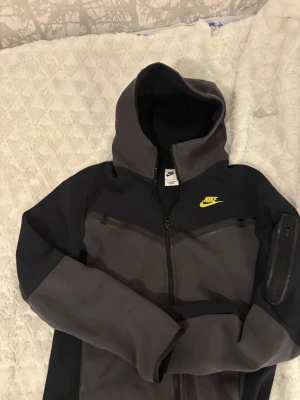 Nike mörkgrå vindjacka med huva - Nike tech i mörkgrå och svart med gul logga på bröstet. Jackan har huva, hel dragkedja och en ficka med dragkedja på ärmen. Snygg, sportig look och slim passform som sitter bra på kroppen.