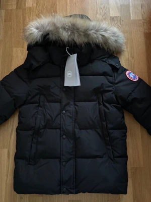 Svart dunjacka med päls från Canada Goose - Säljer en svart dunjacka från Canada Goose med fluffig päls på huvan och klassisk logga på ärmen. Jackan har dragkedja och knappar framtill, två stora fickor och quiltad design. Perfekt för kalla vinterdagar och riktigt snygg stil. Pris går att diskutera.