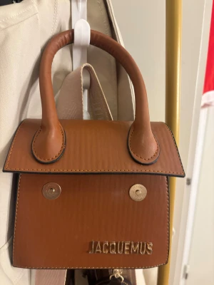 Brun Jacquemus handväska i skinn - Säljer en snygg brun handväska från Jacquemus med stilren design och guldfärgad logotyp framtill. Väskan har två rejäla handtag och magnetknappar på framsidan. Perfekt för dig som gillar trendiga accessoarer med lyxig känsla.