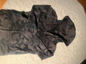 Camouflage hoodie från Polo Ralph Lauren - Säljer en mörkgrå och svart camouflage hoodie från Polo Ralph Lauren. Tröjan har dragkedja, huva med snörning och långa ärmar. Materialet är mjuk bomull och polyester, perfekt för chill dagar. Diskret Polo-logga på bröstet och coolt camomönster över hela tröjan. Använd i några månader, men säljer då den har blivit för liten. Har ett litet hål längst ner vid armen som knappt syns vid användning. Kontakta gärna om du har frågor😊