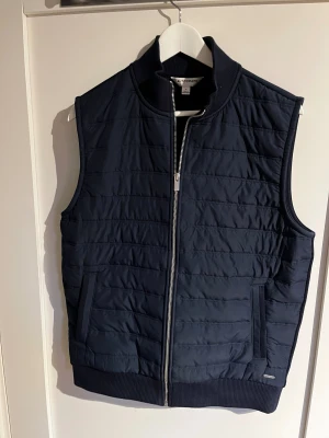 Blå väst från Autograph - Stilren blå väst från Autograph i storlek M. Västen har quiltad framsida, ribbad krage och nederkant, samt praktiska sidofickor. Dragkedja framtill och slät baksida ger en clean look. Perfekt att slänga över en hoodie eller tröja.