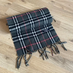 Rutig halsduk från Burberry i ull - Klassisk rutig halsduk från Burberry i 100% lammull. Mörkgrå bas med svarta, vita och röda rutor samt fransar i ändarna. Tillverkad i England och mäter cirka 150 cm. Perfekt accessoar för att lyfta din vinterstil.