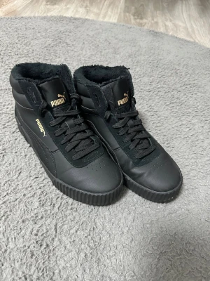 Svarta höga sneakers från Puma - Säljer ett par svarta höga sneakers från Puma med SoftFoam+ innersula för extra komfort. Skorna har pälsfoder, snörning och detaljer i guld. Ovandel i skinn och mocka, platt sula med räfflad kant och rund tå. Perfekta för dig som gillar stilrena och varma sneakers.