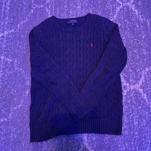 Mörkblå kabelstickad tröja Polo Ralph Lauren - Ordinarie pris 1200 Storlek L14-16, passar dig som är mellan 160-165 .Kabelstickad mörkblå tröja från Polo Ralph Lauren med klassisk rund hals och lång ärm. Tröjan har det ikoniska rosa broderade logot på bröstet och breda ribbade muddar vid ärmslut och nederkant. Perfekt för lager-på-lager under kyliga dagar. Orginal pris 1800