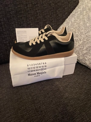 Maison Margiela svarta sneakers - Maison Margiela sneakers i svart skinn med beige detaljer och snörning. Klassisk rund tå och platt gummisula i ljusbrun färg. Snygg minimalistisk design med kontrasterande vita skosnören och innersida. Perfekt för dig som gillar stilrena och exklusiva sneakers. För mer frågor så hör av er👊⚡️