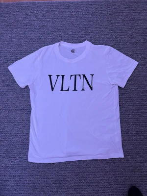 Vit valentino t-shirt  - Vit t-shirt från VLTN i mjuk 100% bomull med klassisk rund hals och svart VLTN-tryck på bröstet. Modellen är kortärmad och har en clean, stilren look som funkar till jeans eller shorts. Perfekt för dig som gillar enkel och snygg streetstil.