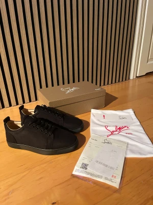 Svarta sneakers från Christian Louboutin - Snygga svarta sneakers från Christian Louboutin med klassisk röd sula och diskret guldfärgad logga på insidan. Skorna har rund tå, snörning och är tillverkade i mocka med svart gummisula. Perfekt för dig som gillar stilrena och lyxiga detaljer.