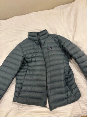 Ljusblå dunjacka från Patagonia XL - Snygg ljusblå dunjacka från Patagonia i storlek XL. Jackan har quiltad design, hög krage och dragkedja framtill. Tillverkad av nylon och fylld med gåsdun och fjädrar. Perfekt för kalla dagar och har en clean, sportig look.