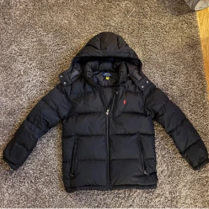 Svart dunjacka från Polo Ralph Lauren - Svart dunjacka från Polo Ralph Lauren med huva och dragkedja framtill. Jackan har två sidofickor med dragkedja och ett litet rött Polo-logotryck på bröstet. Passar L 14-16