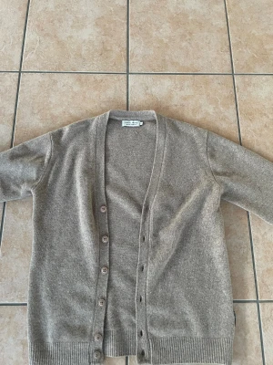 Beige kofta i lammull från Casual Friday - Snygg beige kofta från Casual Friday i mjuk lammull, tillverkad i Italien. Modellen har v-ringning, ribbade muddar och knappar framtill. Perfekt för lager-på-lager och chill stil. Nästan helt oanvänd ! 