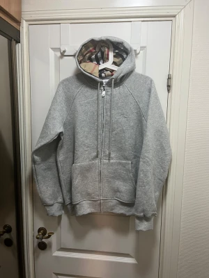 Grå hoodie med dragkedja från Burberry - Säljer en grå hoodie från Burberry med klassiskt rutigt foder i huvan. Tröjan har dragkedja framtill, snörning vid huvan och stora fickor. Mjuk bomullskänsla och ribbade muddar vid ärmslut och nederkant. Perfekt för chill dagar.