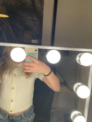 Krämvit stickad blus med guldfärgade knappar - Superfin krämvit stickad blus från Aylin med korta ärmar och snygga guldfärgade knappar framtill. Två dekorativa bröstfickor med knappar ger en chic touch. Ribbad kant vid hals, ärmslut och nederkant. Perfekt till jeans eller kjol!