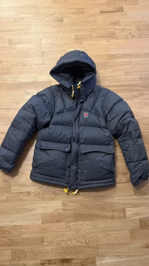Fjällräven Expedition Down Lite - Säljer en mörkgrå Fjällräven Expedition Down Lite med gul snörning och huva. Jackan har dragkedja, knappar och två stora fickor framtill. Perfekt för kalla dagar med sin fluffiga dunfyllning och coola streetstil. Fjällräven-logga på bröstet.