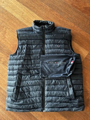 Svart dunväst från Tommy Hilfiger - Snygg svart dunväst från Tommy Hilfiger med quiltad design och ståkrage. Västen har dragkedja framtill, två fickor med knappstängning och ett diskret Hilfiger-logo på bröstet. Lätt och packbar, perfekt för lager-på-lager. Kommer med tillhörande packpåse. Nytt pris för västen är 1350 kr 
