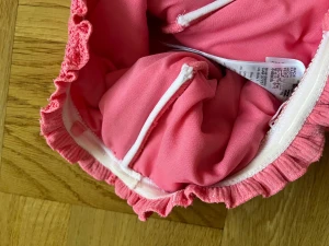 Rosa baddräkt från Billabong - Supersöt rosa baddräkt från Billabong med volangkant upptill och smockad textur. Modellen är axelbandslös och har en tight passform som framhäver figuren. Materialet är mjukt och stretchigt syntet, perfekt för strandhäng och poolparty.