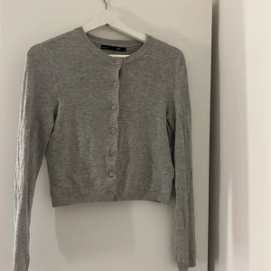 Grå kofta från 157 XS - Grå, ribbad kofta från 157 i storlek XS. Tröjan har rund hals, långa ärmar och knappar hela vägen framtill. Materialet är mjukt och stickat, perfekt för lager-på-lager. Enkel och clean design som funkar till många olika stilar.